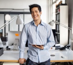 man with a tablet in hand, leaning against his desk and smiling